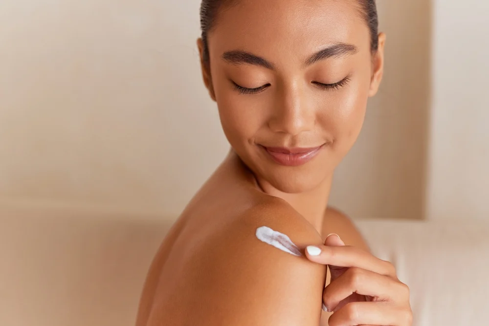 Close-up of a woman looking over her shoulder and applying lotion to tanned skin.