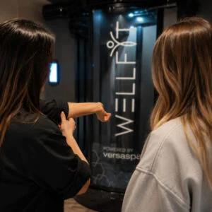 Irish customer showing skincare results after using the WellFit spray-on skincare booth, seen in the background Irish customer showing skincare results after using the WellFit spray-on skincare booth, seen in the background